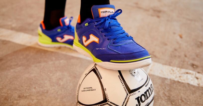 Osoba stojí na futbalovej lopte v halových topánkach značky Joma