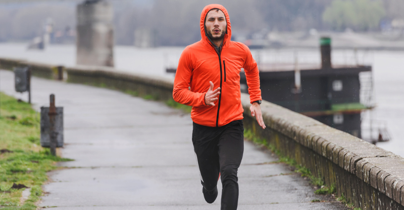 Muž beží po ceste pri rieke počas dažďa v kapucni