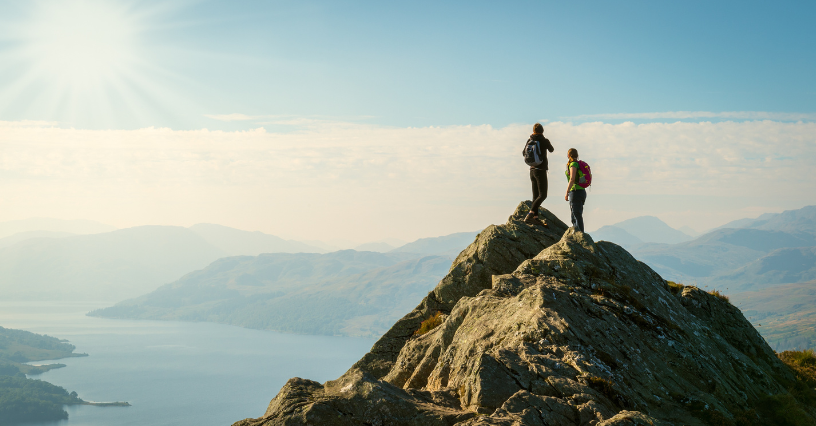 Dve osoby stoja na skalnatom vrchole s výhľadom na jazero, ideálne na prezentáciu turistického vybavenia