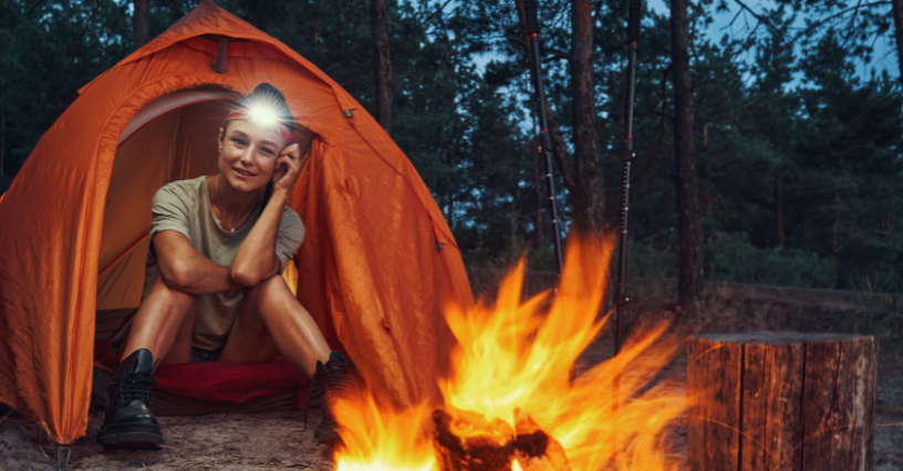 Osoba sedí pred stanom pri ohni a má na hlave čelovku