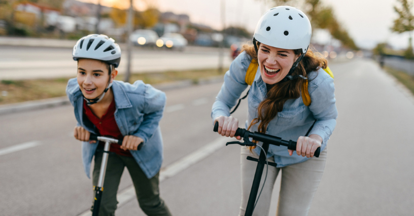 Dve osoby jazdia na kolobežkách po ulici s helmami na hlavách